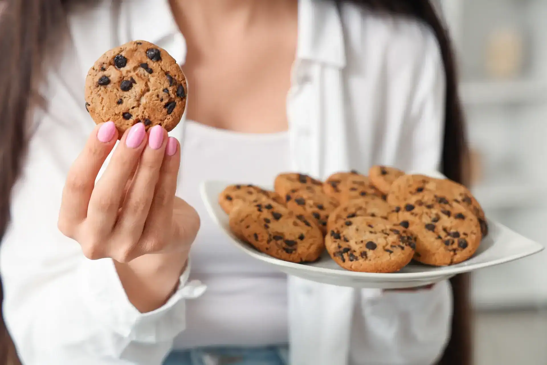 Woman holding a plate of chocolate chip cookies while holding up a cookie in her other hand
